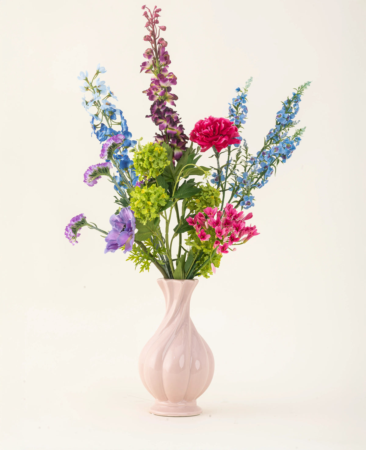 Der Kunstblumenstrauß Blossom Breeze aus Seide ist in einer rosa Keramikvase arrangiert und besteht aus leuchtend blauen, violetten, roten und grünen Blumen – ein wartungsfreier Blumenstrauß vor einem schlichten, hellen Hintergrund.