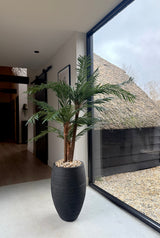 Die künstliche Palme Cycas Classic 135 cm im eleganten Dekotopf wird in der Nähe eines großen Fensters mit Tageslicht platziert. Draußen sieht man ein Strohdach und Kies. Die Inneneinrichtung ist modern und minimalistisch.