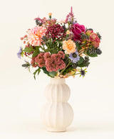 In einer weißen, geschwungenen Keramikvase befindet sich der Kunstblumenstrauß Pretty Pastel aus Seide – eine farbenfrohe Mischung aus nachhaltigen Blumen wie rosa Rosen, Dahlien, lila und orangefarbenen Blumen und Grün – wunderschön arrangiert vor einem hellen Hintergrund.