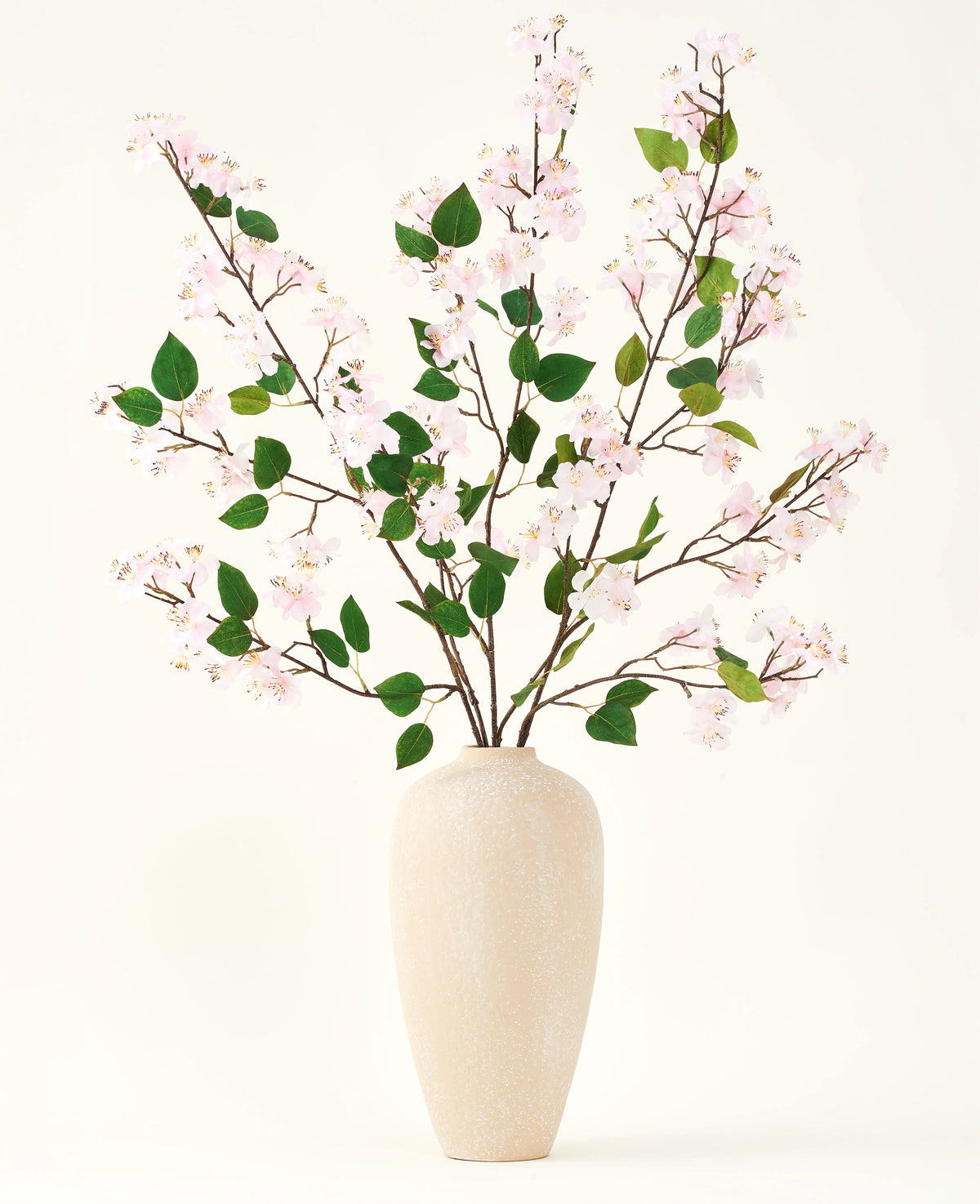 Eine beige Keramikvase enthält mehrere lange Zweige mit dem künstlichen Seidenblumenstrauß aus Pfirsichblüten-Rosa und grünen Blättern vor einem schlichten weißen Hintergrund – perfekt für ein Interieur mit künstlichen Blumen.