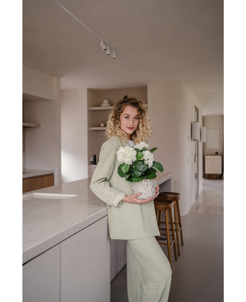 Een vrouw met krullend blond haar, gekleed in een lichtgroene outfit, staat in een moderne keuken met een Kunstplant Hortensia Wit UV 35 cm in haar handen – met crèmewitte bloemen – en glimlacht naar de camera.