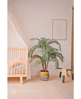 Ein gemütliches Kinderzimmer mit einem hellen Holzbettgestell, der künstlichen Palme Cycas Classic 135 cm in einem gelben Pflanzgefäß, einem hölzernen Spielzeugfahrrad mit der Aufschrift MIA, minimalistischer Strichzeichnung an der Wand, hellen Holzböden und zartrosa Bettwäsche für zusätzliche Wärme.