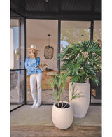 Eine Frau mit Hut, blauer Bluse und weißer Hose lehnt an einer Glastür. Im Vordergrund steht die Kunstpflanze Philodendron 125 cm. Im Inneren erwartet Sie ein gemütlicher, moderner Wohnraum.