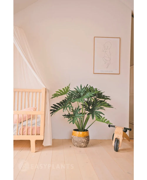 Ein gemütliches, minimalistisches Kinderzimmer mit einer hölzernen Wiege und einem Baldachin links, einer künstlichen Philodendron-UV-Pflanze 90 cm als Prunkstück in der Mitte, rechts einem hölzernen Spielzeugfahrrad und darüber einem einfachen Line-Art-Druck.