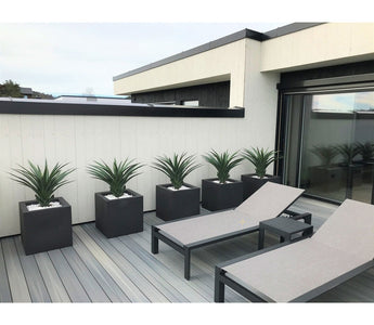 Auf einer modernen Terrasse stehen zwei graue Liegestühle und fünf quadratische Pflanzgefäße, jeweils mit einer künstlichen Pflanze Agave Pandora UV 85 cm, vor einer weiß-schwarz getäfelten Wand für einen eleganten, minimalistischen Look.