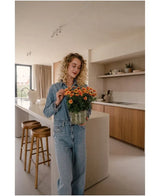 Eine Frau mit lockigen blonden Haaren, die ein Jeanshemd und Jeans trägt, steht in einer modernen Küche und hält in ihren Händen eine künstliche Gänseblümchen-UV-Pflanze von 31 cm mit leuchtend orangefarbenen Blüten. Neben ihr stehen zwei Holzhocker auf der Kücheninsel.