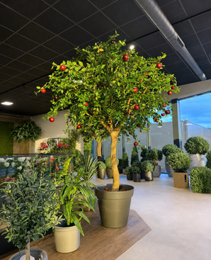 Kunstappelboom van 240 cm met levensechte rode appels in een bruine pot, omringd door diverse groene potplanten in een moderne, goed verlichte showroom met glazen deuren en ramen. Dit indrukwekkende kunstboom-interieurstuk geeft elke ruimte een levendige, natuurlijke uitstraling.