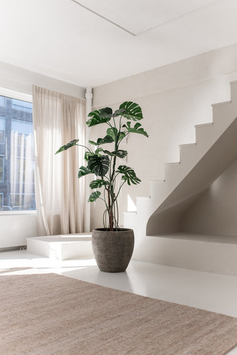 Die Kunstpflanze Monstera 210 cm steht in einem Topf auf einem weißen Boden neben einer modernen Treppe in einem hellen, minimalistischen Raum, in den natürliches Licht durch beige Vorhänge fällt.