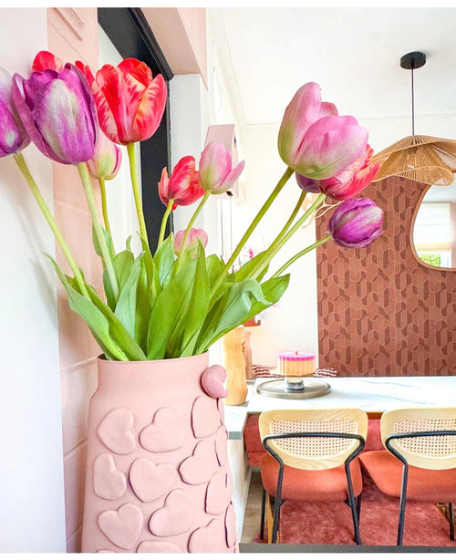 Eine rosa Vase mit geprägten Herzen hält die Kunstblume Französische Tulpe 60 cm rot auf einem Esstisch. Dahinter stehen Stühle mit Korbrücken, ein Kuchen auf dem Tisch und ein Spiegel an einer gemusterten braunen Wand unter einer geflochtenen Hängelampe.