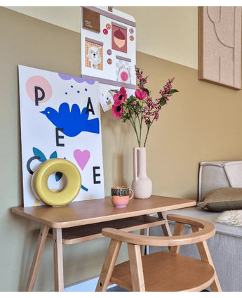 Ein kleiner Holzschreibtisch mit Stuhl ist mit künstlicher Katzenminze 75 cm rosa, einer Tasse, einer gelben Ringskulptur und bunten Kunstdrucken an der Wand darüber dekoriert. Rechts ist teilweise eine Bank zu sehen.