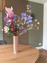 Eine hohe rosa-weiß gestreifte Vase mit dem künstlichen Seidenblumenstrauß Amethyst Romance steht auf einem runden Holztisch in einem modernen Raum mit grünen Wänden, Holzvertäfelung und kleinen gerahmten Kunstwerken.