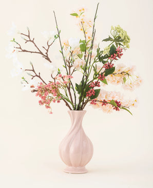 Een roze vaas van keramiek bevat het Zijden kunstbloemen boeket Springtime Poem, met witte, roze en groene bloemen met groene bladeren en bruine stelen, tegen een lichte achtergrond.