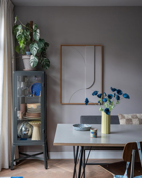 Der moderne Essbereich verfügt über eine graue Glasvitrine, in der Geschirr und Pflanzen ausgestellt sind, minimalistische Wanddekorationen und einen Tisch, auf dem die Kunstblume Mohn 73 cm dunkelblau in einer gelben Vase steht. Abgerundet wird der Raum durch eine eklektische Mischung aus Stühlen, alle vor dem Hintergrund grauer Wände und eines Holzbodens.
