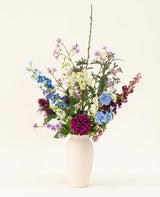 Eine weiße Vase mit dem künstlichen Seidenblumenstrauß „Purple Rain“ zeigt leuchtend violette und weiße Kunstblumen und grüne Blätter, ein Beispiel niederländischer Handwerkskunst vor einem hellen Hintergrund.