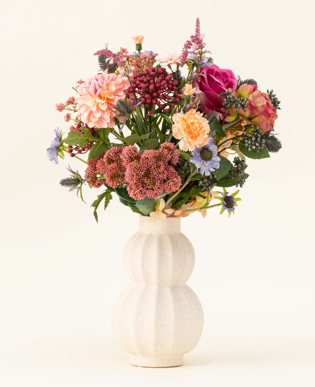 In einer weißen, geschwungenen Keramikvase befindet sich der Kunstblumenstrauß Pretty Pastel aus Seide – eine farbenfrohe Mischung aus nachhaltigen Blumen wie rosa Rosen, Dahlien, lila und orangefarbenen Blumen und Grün – wunderschön arrangiert vor einem hellen Hintergrund.