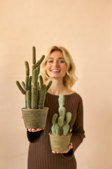 Eine lächelnde Frau in einem braunen Kleid hält zwei künstliche Kaktus-Cereus-Topfpflanzen (35 cm), eine große und eine kleine, vor einem schlichten beigen Hintergrund.