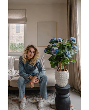Een vrouw met krullend blond haar, gekleed in een spijkeroutfit, zit op een bank naast de Kunstplant Hortensia UV 85 cm blauw, een hoge witte vaas met blauwe hortensia’s, in een moderne, lichte woonkamer.
