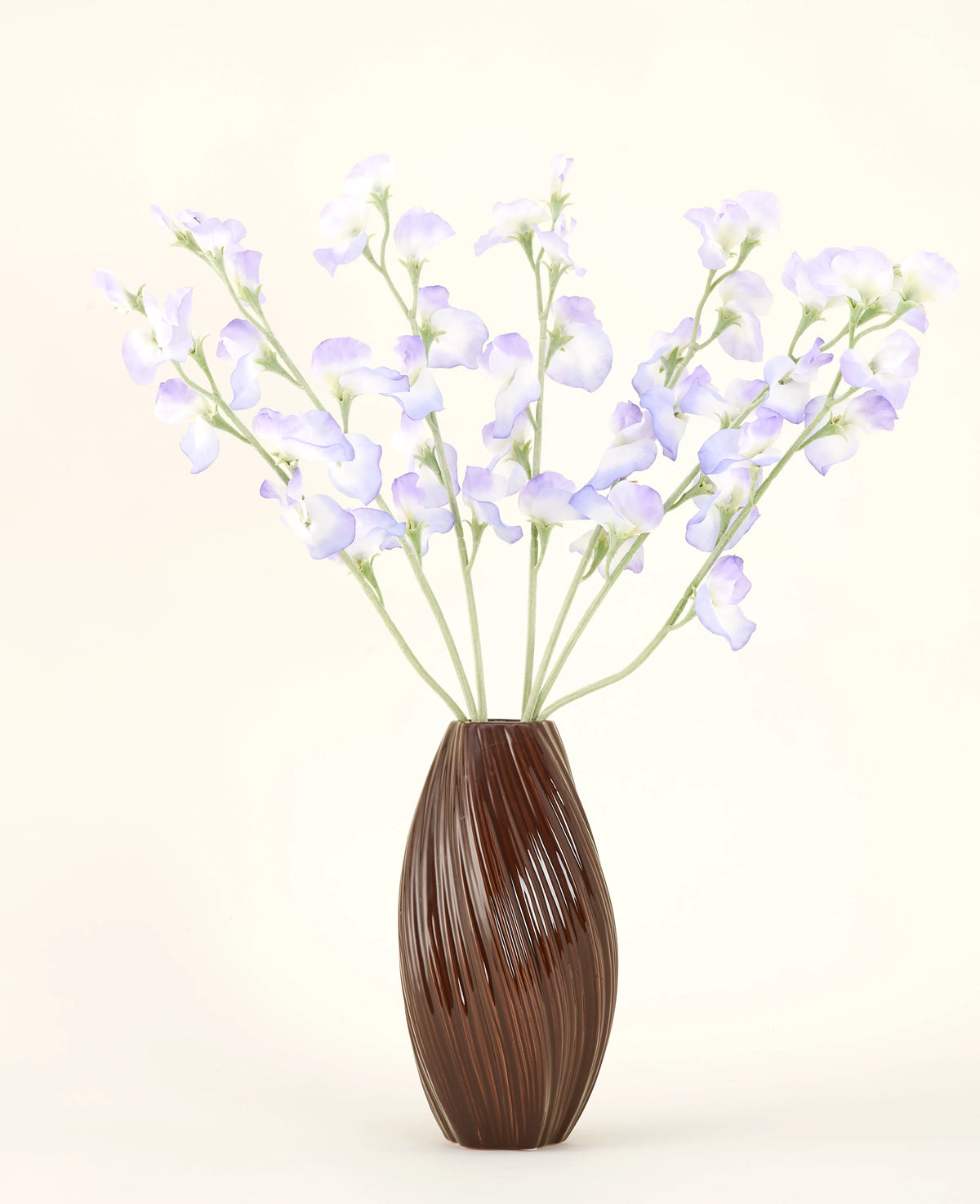 In einer braunen, gerippten Vase befindet sich der künstliche Seidenblumenstrauß Erbsen-Lavendel mit zarten Blütenblättern und grünen Stielen vor einem schlichten, cremefarbenen Hintergrund für eine dezente Landhausatmosphäre.