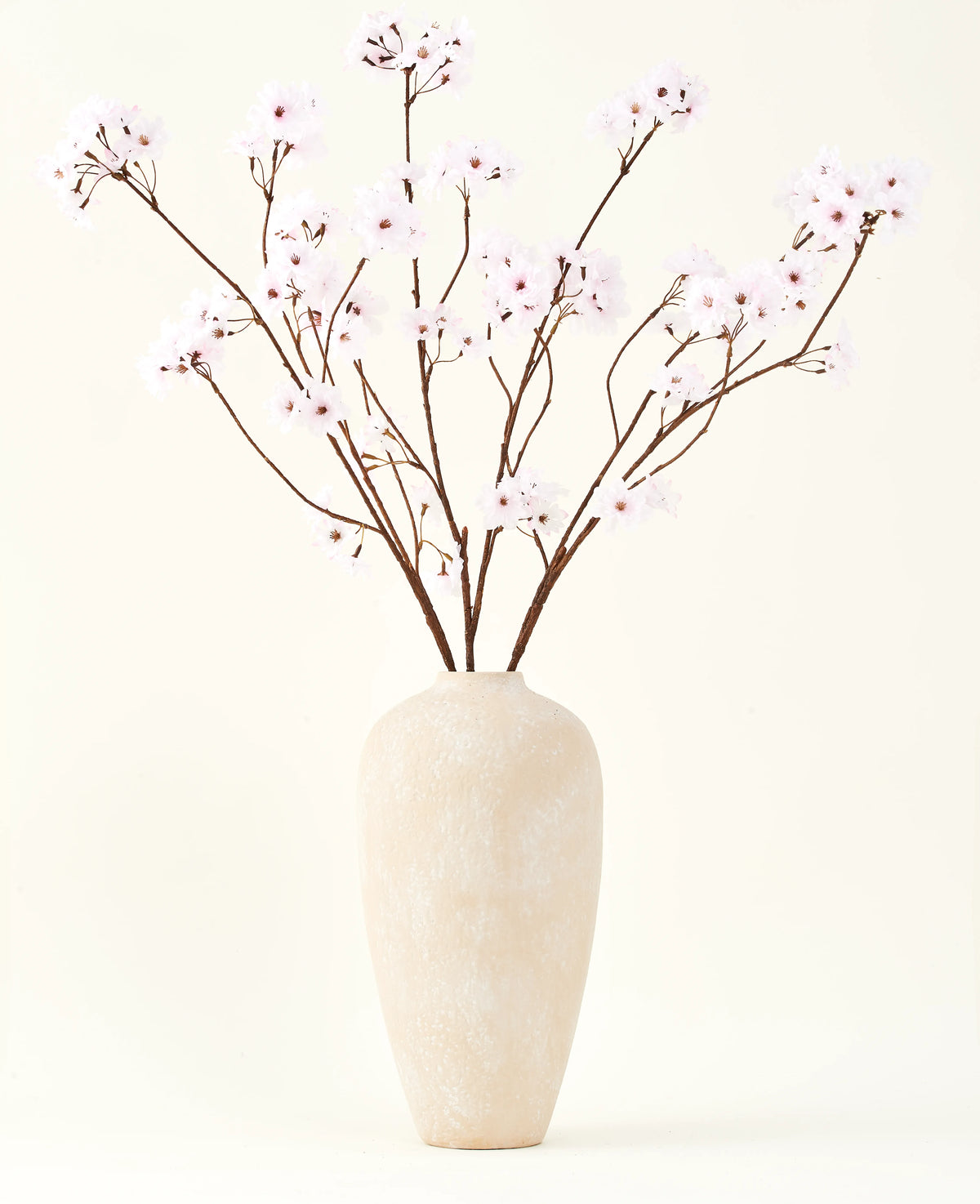 Der Seiden-Kunstblumenstrauß Kirschblüten hellrosa 114 cm steht in einer hohen beigen Keramikvase mit langen, dünnen Zweigen und zarten hellrosa Blüten und setzt einen ruhigen, künstlichen Blumenakzent vor einem schlichten Hintergrund.