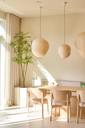 Een lichte, minimalistische eethoek is ingericht met stoelen van licht hout, een tafel, drie papieren hanglampen, de Kunstplant Mahonia van 210 cm in een pot en gordijnen die van de vloer tot aan het plafond reiken bij een groot raam dat zonlicht binnenlaat.