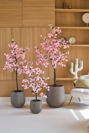Drie potplanten van het type Kunstplant Bougainvillea van 150 cm, in roze met roze bloemen, staan opgesteld voor houten planken, naast een moderne witte stoel met een kussen en een decoratieve kandelaar.