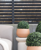 Drie Kunstplant Buxus Romeinse Stijl UV D30cm ronde topiaries staan op een patio bij een beige buitenstoel, tegen een modern zwart latten hek en kale bomen.