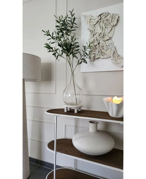 Een minimalistische consoletafel toont een heldere vaas met de Kunst Olijftak 109 cm, een ronde vaas van wit keramiek en een witte schaal met LED-kaarsen. Erboven hangt gestructureerde witte wandkunst en vlakbij staat een staande lamp.