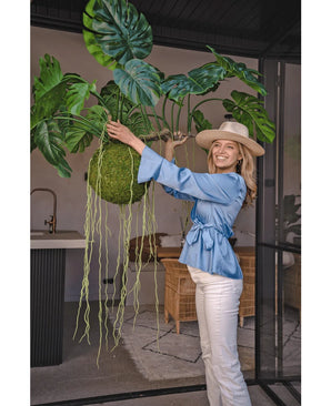 Een glimlachende vrouw in een blauwe blouse, witte broek en hoed toont de Kunst hangplant Monstera Deluxe 80 cm met slingerende groene wijnranken, die een tropische sfeer brengt in de lichte binnenruimte met glazen wanden en rotan meubilair.