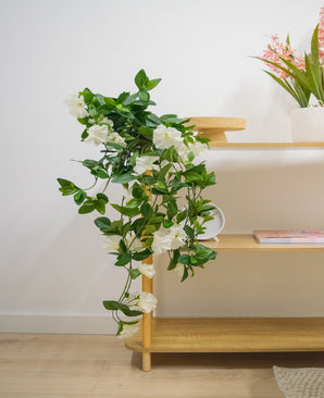 Die künstliche Hängepflanze Petunie 70 cm weiß mit weißen Blüten fällt von einem Holzregal in einen hellen, modernen Raum mit hellen Holzböden. Auf dem obersten Regal steht eine Vase mit langen rosa Blumen.