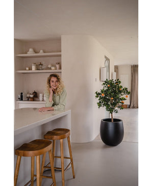 Eine Frau mit blonden Locken lächelt und lehnt an einer Theke. Neben ihr sorgen zwei Holzhocker und ein künstlicher Orangenbaum 120 cm für eine frische Atmosphäre in der modernen, hellen Küche mit Regalen und Tellern dahinter.