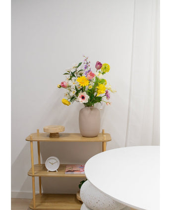Een lichthouten plank toont een beige vaas met Kunstranonkel Real Touch Geel 60cm bloemen, een witte klok en een houten dienblad. Een deel van een ronde witte tafel verschijnt op de voorgrond tegen een effen witte muur.