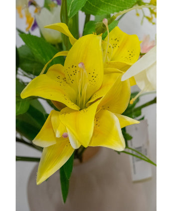 Een close-up van de Kunstbloem Aziatische Lelie 66 cm geel, met heldergele bloemblaadjes met donkere spikkels, omringd door groene bladeren en andere bloemen in een lichtgekleurde vaas.