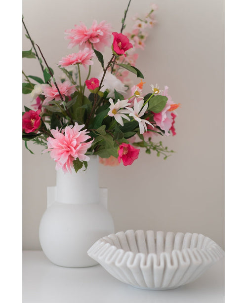 Eine Kunstblumen-Dahlie 68 cm rosa in einer weißen Vase steht neben einer weißen Keramikschale mit Rüschen auf einer weißen Fläche vor einem schlichten hellen Hintergrund.