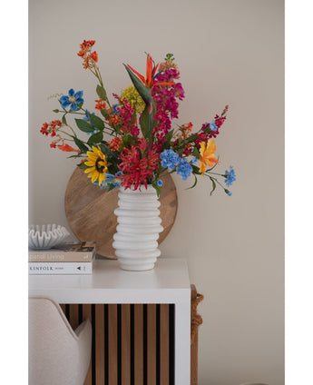 Auf einem weißen Tisch neben gestapelten Büchern und Deko steht in einer weißen, strukturierten Vase ein 75 cm großer künstlicher Blumenstrauß aus Katzenminze in Rosa, im Hintergrund lehnt ein rundes Holztablett an der beigen Wand.