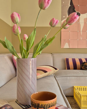 Een Kunstbloem Franse Tulp van 63 cm in roze staat in een vaas op de tafel naast een oranje kopje in een gezellige woonkamer met een crèmekleurige bank, kussens met patronen, een gele krat en abstracte wandkunst op de achtergrond.