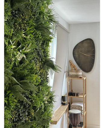 Een weelderige Kunsthaag Freshness 100x100 cm UV verticale tuin bedekt één muur van een lichte kamer. Vlakbij hangt een moderne driehoekige wandklok boven een houten plank met handdoeken, een djembé trommel, manden en kleine decoratieve stukken.
