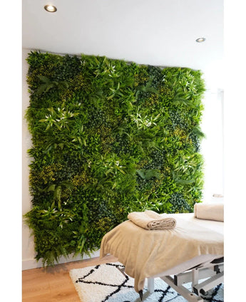 Een massagetafel met opgevouwen handdoeken staat voor een Kunsthaag Freshness 100x100 cm UV verticale tuin in een lichte, moderne kamer met witte muren en een geometrisch tapijt.