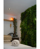 Een rustige spa-ruimte met een massagetafel, rustgevende verlichting, een Himalayazoutlamp op een plank en een weelderige Kunsthaag Freshness 100x100 cm UV groene plantenwand voor extra sereniteit.