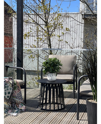 Een modern terras met twee stoelen met kussens, een ronde zwarte tafel met de Kunstplant Laurier UV d33 cm als middelpunt en een deken met patroon op één stoel. Bomen en een houten hek verschijnen op de achtergrond onder een heldere hemel.
