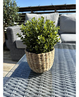 De Kunstplant Buxus groen in pot UV 19 cm, met groene bladeren in een rieten mandvormige pot, staat op een glazen buitentafel met terrasmeubilair en een houten reling zichtbaar onder een strakblauwe hemel.