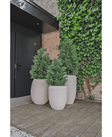 Drie grote beige plantenbakken met Kunstplant Conifeer deluxe UV 120 cm struiken staan op een houten dek bij een zwarte deur en een met klimop begroeide muur en bieden UV-bescherming voor blijvende schoonheid in je kunstplant tuin.