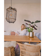 Een vrouw in een witte blouse zit aan een houten eettafel met rieten stoelen. De gezellige, zacht verlichte kamer heeft de Kunstplant Alocasia UV 67 cm en een hanglamp met textuur als decor.