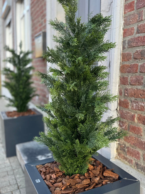 Eine Nahaufnahme der künstlichen Nadelbaumpflanze UV ​​120 cm, einer grünen künstlichen Nadelbaumpflanze in einem rechteckigen schwarzen Pflanzgefäß mit braunem Rindenmulch, ausgestellt auf einem Bürgersteig in der Nähe eines Backsteingebäudes; Ein weiterer solcher Pflanzer ist im Hintergrund.