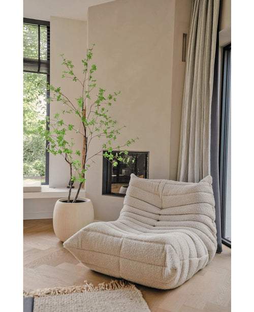 Ein gemütlicher, cremefarbener, strukturierter Loungesessel steht neben einem großen Fenster und einem Kamin. In der Nähe verleiht die künstliche Enkianthus-Pflanze (180 cm) dem hellen, luftigen und modernen Raum einen natürlichen Hauch von Grün.