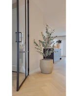 Een modern interieur met lichte houten vloeren, een grote glazen deur en de Kunstplant Eucalyptus 150 cm. Op de achtergrond staat een donkere kast terwijl zacht natuurlijk licht door de gordijnen naar binnen valt.