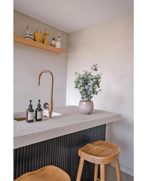 Auf einer modernen Arbeitsplatte befinden sich ein Wasserhahn aus Messing, Seifenspender, die künstliche Eukalyptus-Topfpflanze (61 cm) und zwei Holzhocker. Über der Arbeitsplatte befinden sich Regale an einer hellen Wand, auf denen Gläser und Flaschen Platz finden.