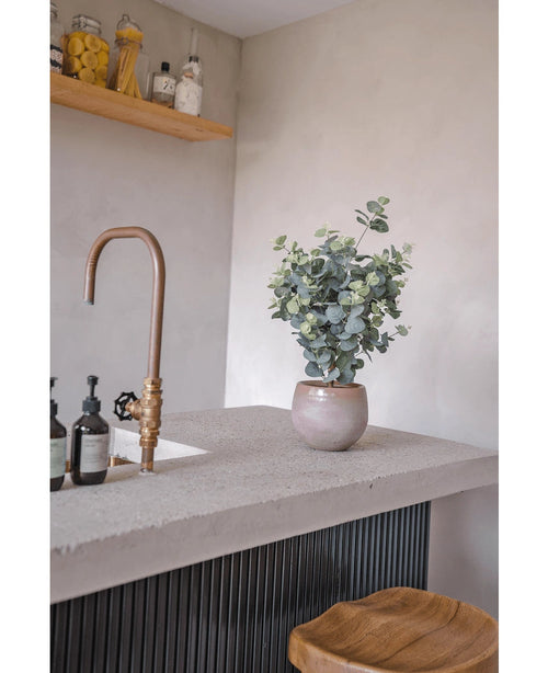 Eine moderne Arbeitsplatte mit einem Wasserhahn aus Bronze, Seifenflaschen, einem Holzhocker und dem künstlichen Eukalyptus 76 cm. Im Hintergrund sind an einer hellgrauen Wand Holzregale mit Gläsern und Flaschen montiert.
