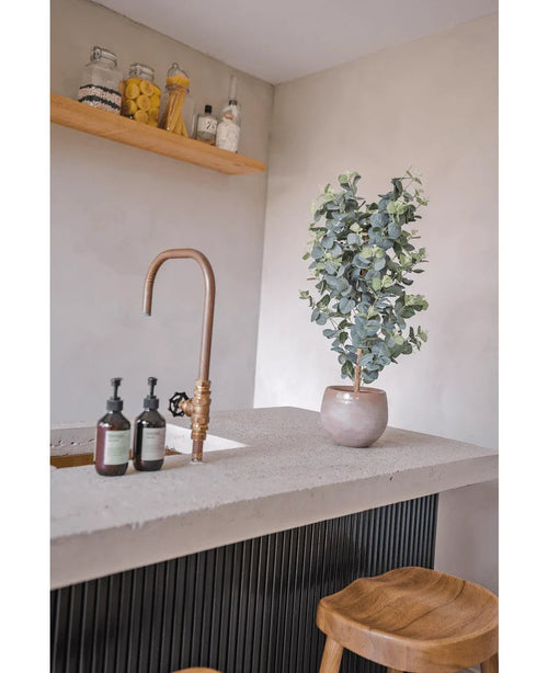 Eine moderne Arbeitsplatte verfügt über einen Messinghahn, Seifenspender, Holzhocker und die künstliche Eukalyptuspflanze 91 cm. Gläser mit Vorratsutensilien stehen auf einem Holzregal vor einer hellen Wand.
