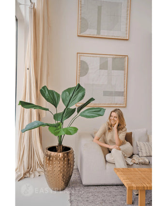 Een vrouw ontspant op een lichte bank in een moderne, neutraal gekleurde woonkamer. Naast haar staat de Kunstplant Lucuala Grandis 120 cm in een pot, terwijl er abstracte kunst boven hangt en natuurlijk licht de ruimte verlicht.