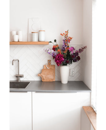 Eine moderne weiße Küche mit einer grauen Arbeitsplatte, einer Spüle, einem Holzschneidebrett und einer weißen Vase gefüllt mit einer künstlichen Hortensie 55 cm in Dunkelrosa. Auf einem Holzregal über der Theke stehen weiße Tassen und Töpfe.